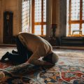 Man in a white prayer cap kneels in prostration on a richly patterned carpet inside a sunlit room, likely a mosque.
