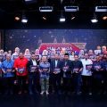 Group of people on a stage holding certificates, posing for a photo at an event with a decorative backdrop and banner behind them