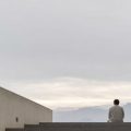 Person seated on wide concrete steps, facing a distant misty mountain range between tall, modern buildings on either side.