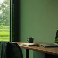 Work desk by a green wall and large window, with laptop, notebook, mug, and books, plus a potted plant beside the screen.