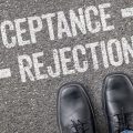 Feet in black dress shoes stand on asphalt beneath large white arrows and the words 'Acceptance' and 'Rejection'.