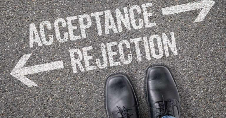Feet in black dress shoes stand on asphalt beneath large white arrows and the words 'Acceptance' and 'Rejection'.