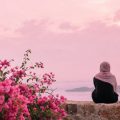 Woman wearing a beige hijab sits with her back to the camera on a stone wall, looking at a pink sunset over the water.