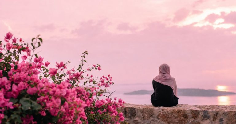 Woman wearing a beige hijab sits with her back to the camera on a stone wall, looking at a pink sunset over the water.