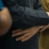 People standing in a line, foreground figure with hands clasped at the waist.”