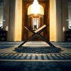 Open Quran stand (rehal) with an open book in a mosque, centered on a patterned prayer rug with warm hanging lights overhead