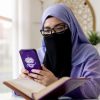 Woman wearing a purple hijab and black face covering, glasses on, looking at a purple smartphone while a book lies open in front of her in a bright, cozy study space.