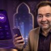 Man in a beige blazer smiles at the camera while holding a smartphone; a floating purple phone interface and a small white robot hover beside him in a techy, glowing set.