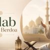 Pria Muslim berjubah putih berdoa dengan tangan terangkat di teras berornamen, mushaf terbuka di depan, latar masjid berkehijauan dan lampu hias di lantai