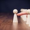 A wooden pawn at the front is touched by a finger, with a line of pawns behind it on a wooden table.
