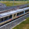Modern silver-beige rapidKL passenger train on elevated tracks, curving rail lines in the background and green fields around the station area.