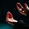 Two hands cupped together in a prayer-like gesture against a dark background with teal light at the bottom left corner.